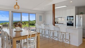 A modern kitchen with white cabinetry, a stainless steel refrigerator, and a view of the ocean through large windows. - Berry's Beach Retreat Ventnor Phillip Island (Ventnor)