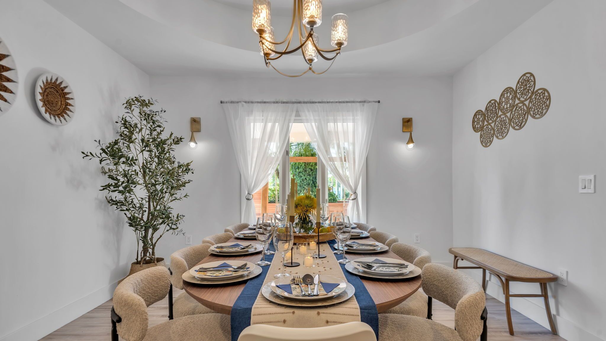 Une salle à manger avec une table ronde dressée pour six personnes, entourée de fauteuils confortables, d’un lustre et offrant une vue à travers de fins rideaux.