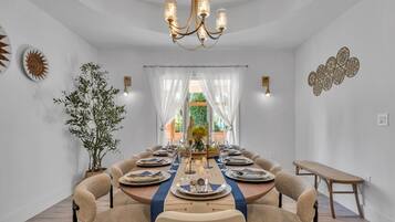 Une salle à manger avec une table ronde dressée pour six personnes, entourée de fauteuils confortables, d’un lustre et offrant une vue à travers de fins rideaux.