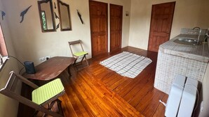 A compact room with wooden flooring, a dining table, two wooden doors, a small kitchenette, and a ceiling fan.