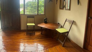 A room with a wooden floor, a table, two chairs, a window with a view of greenery, and wall-mounted mirrors.