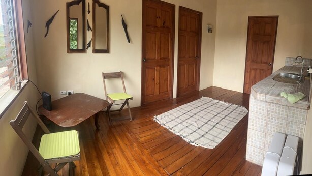 A compact room with wooden flooring, a small table with a chair, a checkered rug, and a kitchenette with a sink and tiled backsplash.