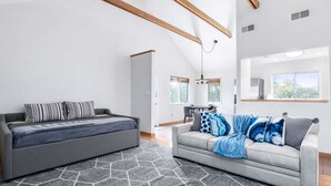Ein modernes Wohnzimmer mit einem grauen Sofa und einer grauen Couch, beide mit gemusterten Kissen. Auf dem Boden liegt ein geometrischer Teppich, ein Deckenventilator und ein Fenster sorgen für natürliches Licht.