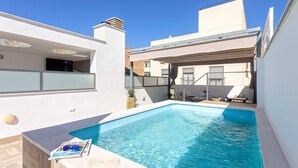 Une piscine avec un magazine posé sur le bord, un espace salon couvert et un bâtiment blanc doté d’un toit plat.