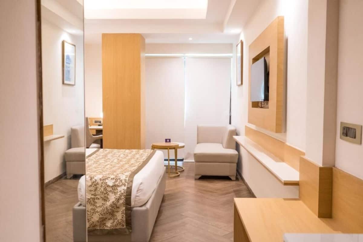 A hotel room with a bed, a desk, a chair, a small table, and a TV mounted on the wall.