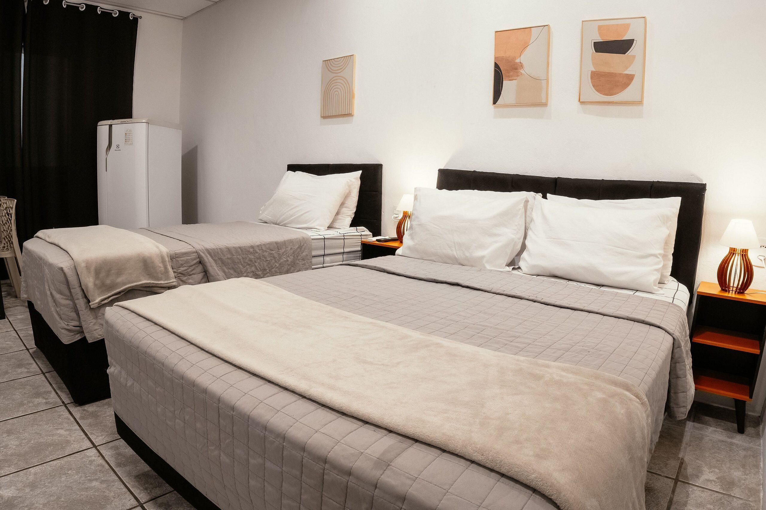 A hotel room with two beds, a refrigerator, a nightstand with a lamp, and abstract wall art.