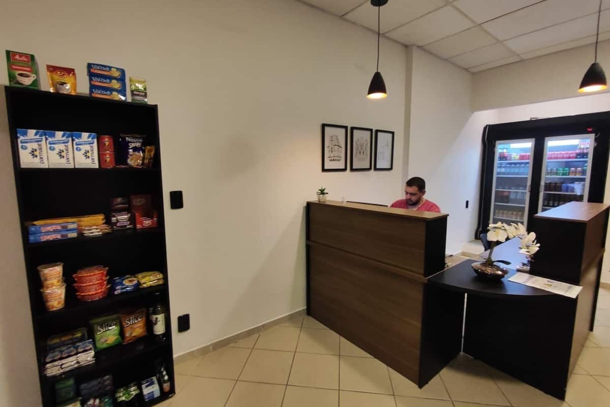 Uma área de recepção com uma mesa de madeira, uma estante com lanches e uma geladeira ao fundo.