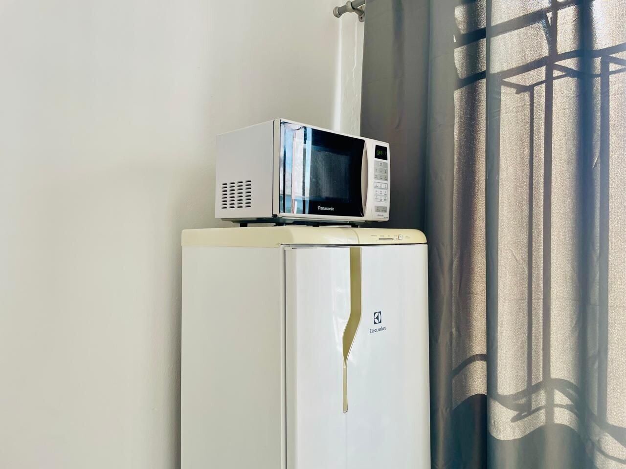 Un refrigerador blanco con un microondas Panasonic encima, un logotipo de Electrolux visible y una ventana con cortina.