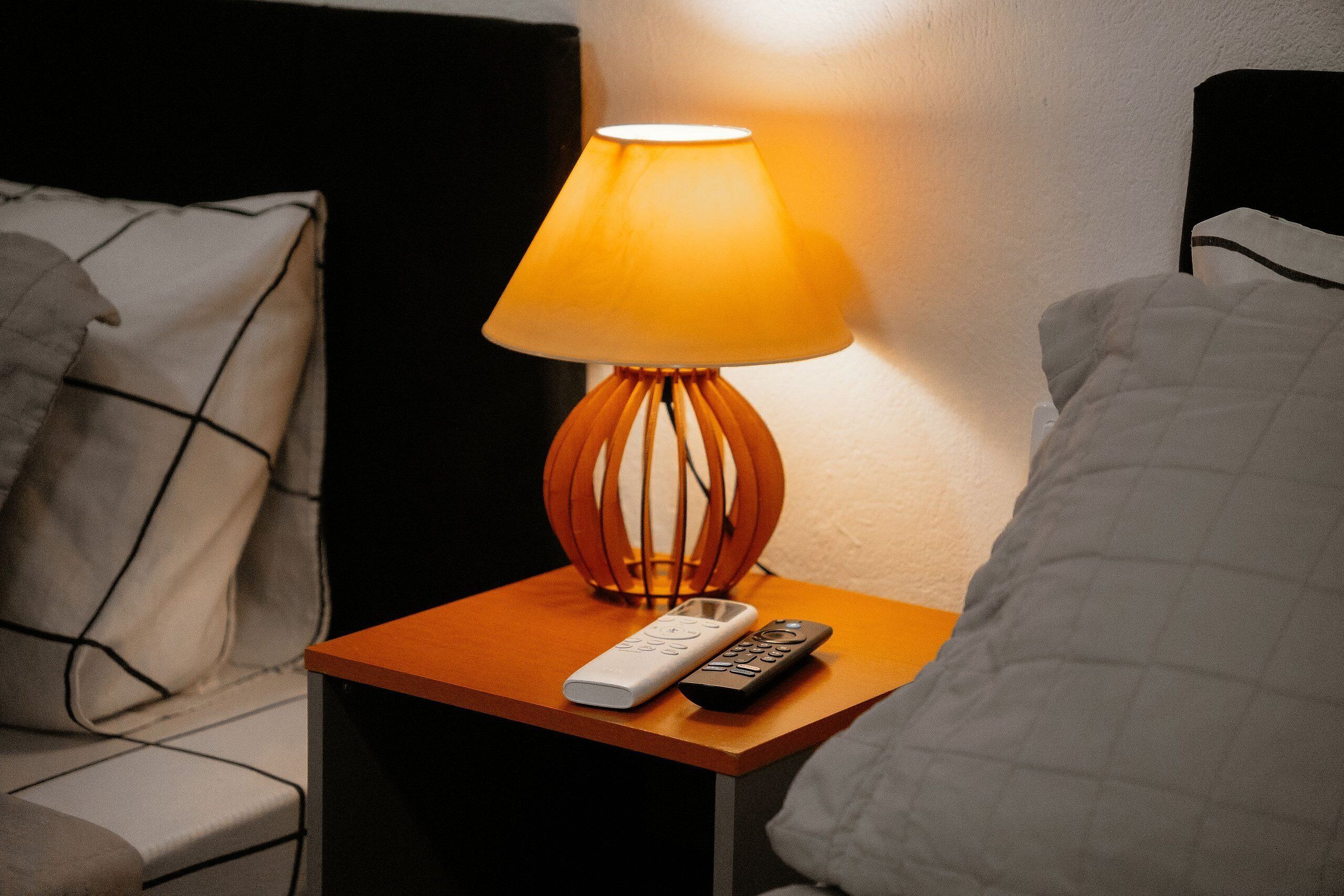 Uma mesa de cabeceira com uma luminária, um controle remoto e um travesseiro.