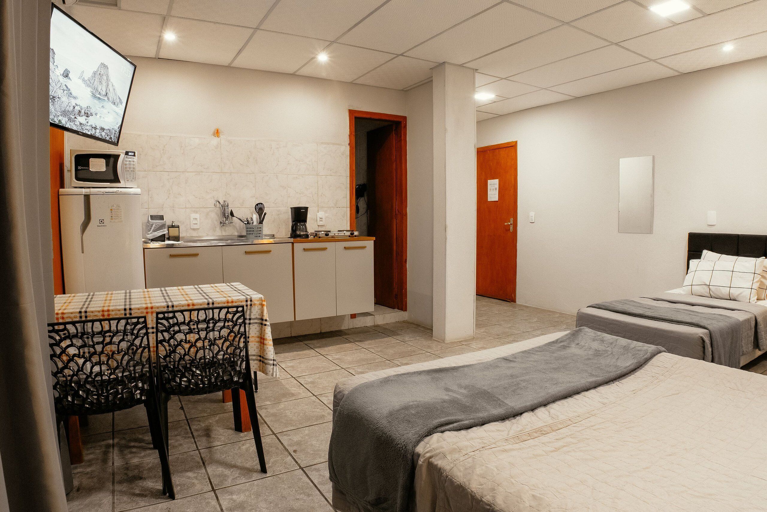 Habitación de hotel con kitchenette, una mesa de comedor con sillas, una cama con manta gris y un televisor montado en la pared.