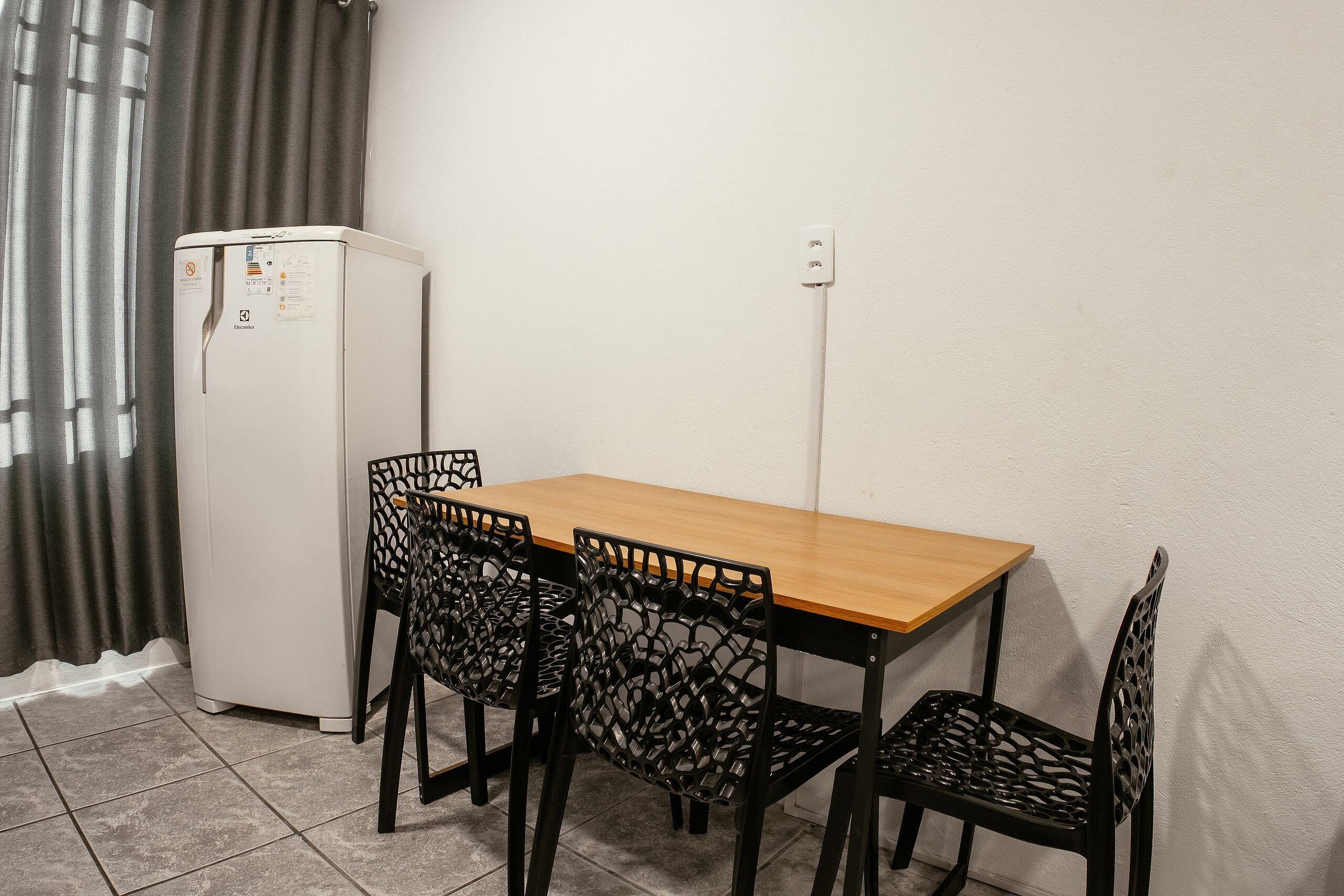 Quarto pequeno com uma mesa de madeira, quatro cadeiras pretas com design vazado, uma geladeira branca e uma tomada na parede.
