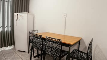 Una habitación pequeña con una mesa de madera, cuatro sillas negras con diseño de celosía, un refrigerador blanco y un enchufe en la pared.
