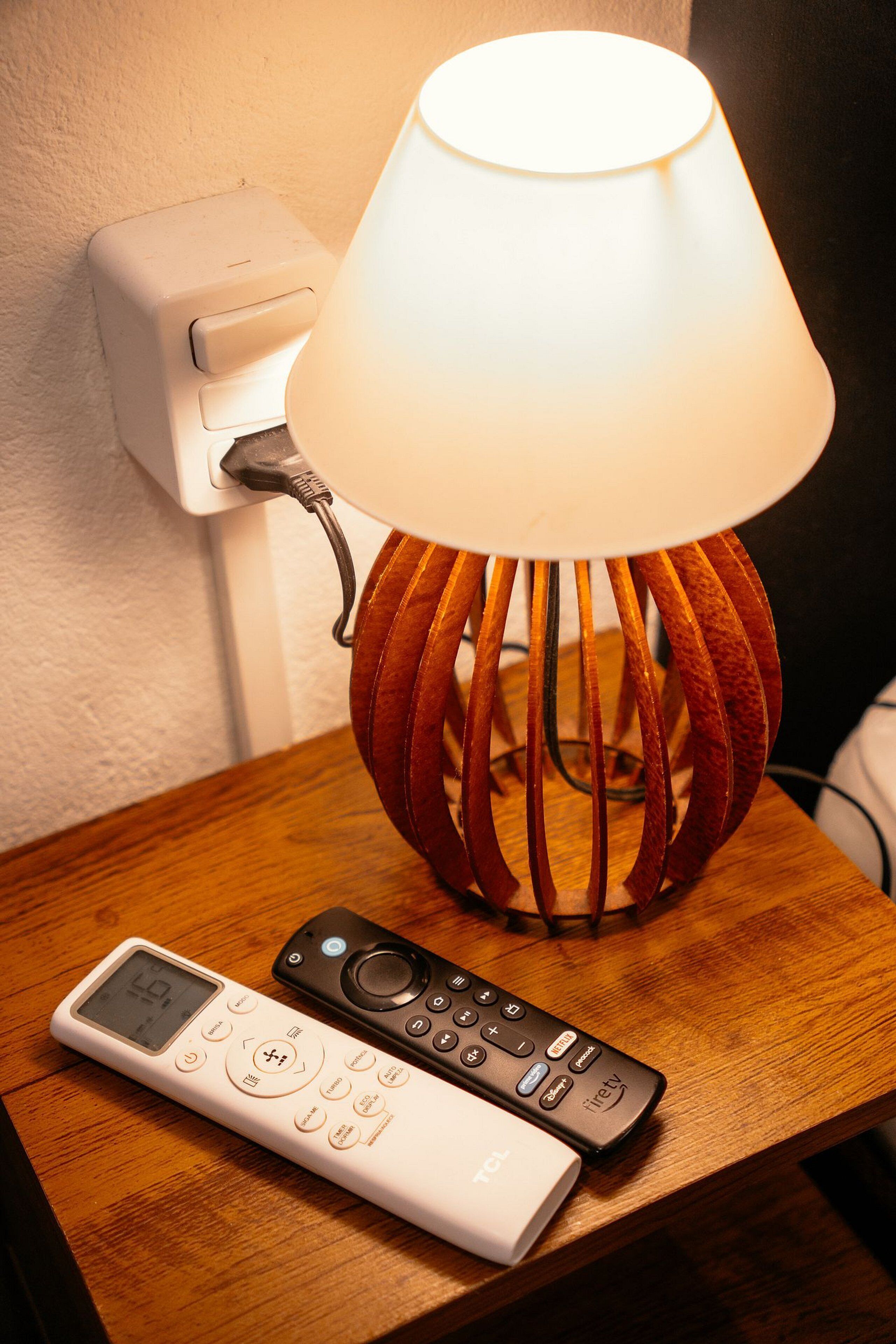 A lamp with a wooden base and a white shade, a remote control, and a power outlet on a wooden surface.