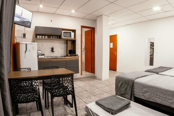 A compact hotel room with a kitchenette, a dining area with a table and chairs, a bed with a gray blanket, and a TV mounted on the wall.