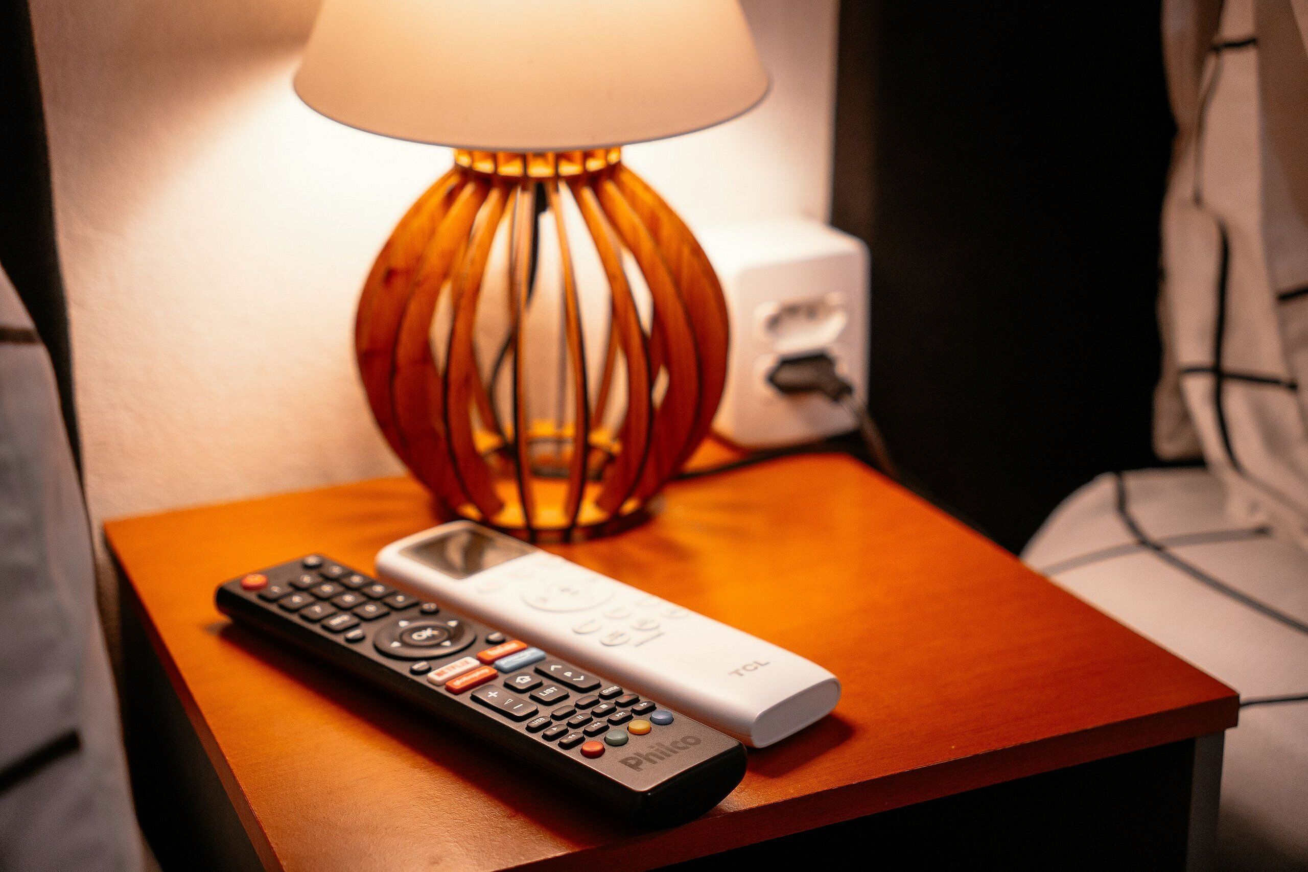 A remote control and a white device on a wooden bedside table with a lamp in the background.