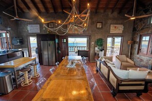 A spacious kitchen with a long wooden table, a refrigerator, and a dining area with a view of the sea.