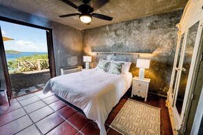 Un dormitorio con una cama, dos mesitas de noche con lámparas, un ventilador de techo, un espejo grande y vista al mar.
