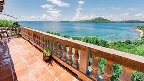 A balcony with terracotta tiles, a potted plant, and a view of a calm sea and green hills.