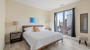 A modern bedroom with a large bed, a nightstand, a lamp, a window with curtains, and a city view. - Fabulous 2BR ! Free Parking !  30 Min to NYC ! (Newark)