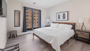 A modern hotel room with a large bed, a bedside table with a lamp, a small stool, and a large window with curtains. - Marvelous 2BR ! Free Parking !  30 Min to NYC (Newark)