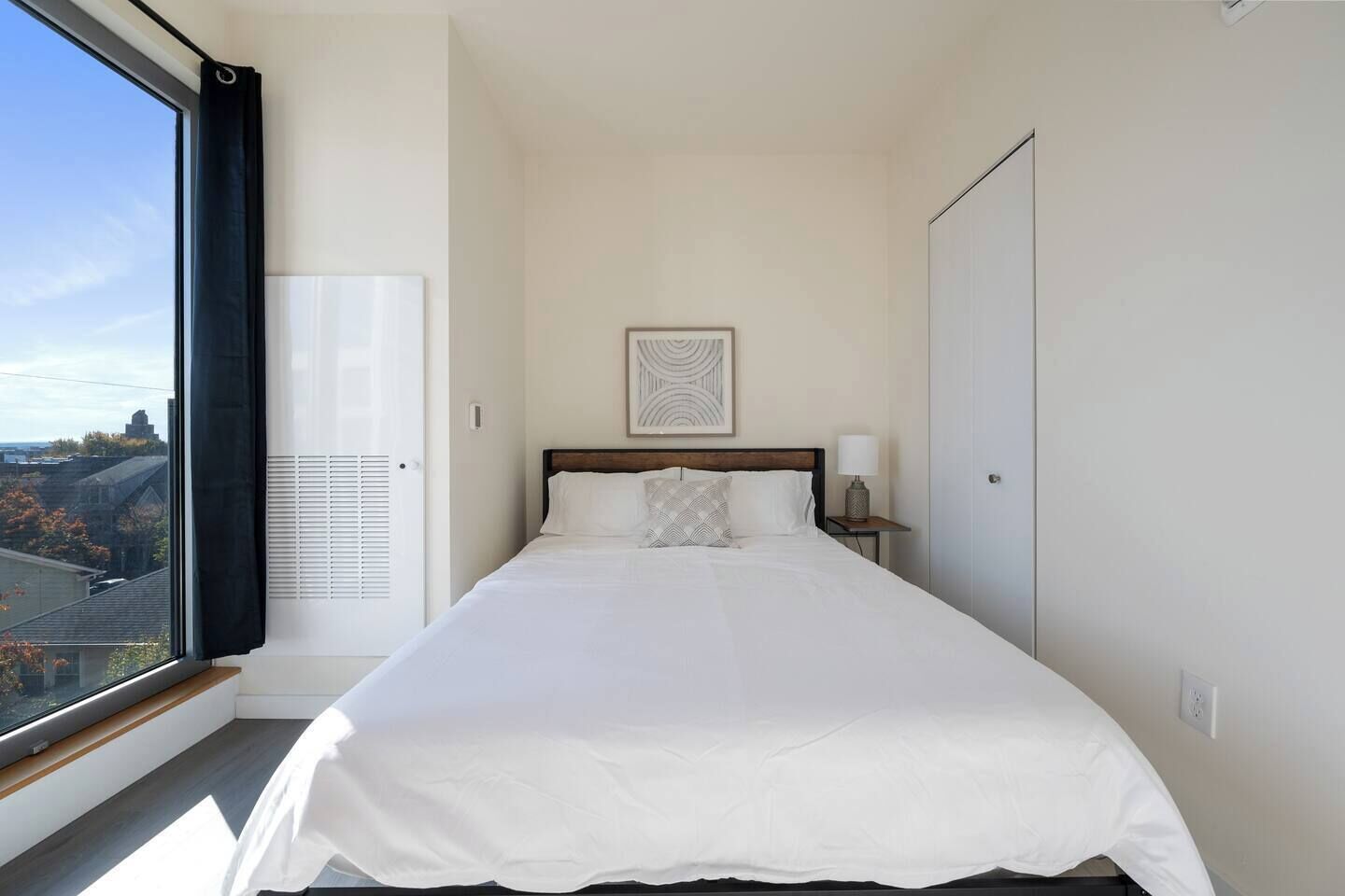 Un dormitorio con una cama grande, una mesita de noche con lámpara, una ventana con vista a edificios y una puerta.