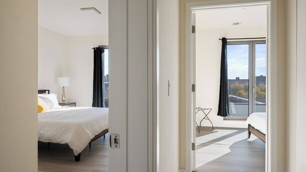 Een moderne hotelkamer met een groot bed, een nachtkastje met een lamp, uitzicht op de stad door een groot raam en een balkon met een stoel.