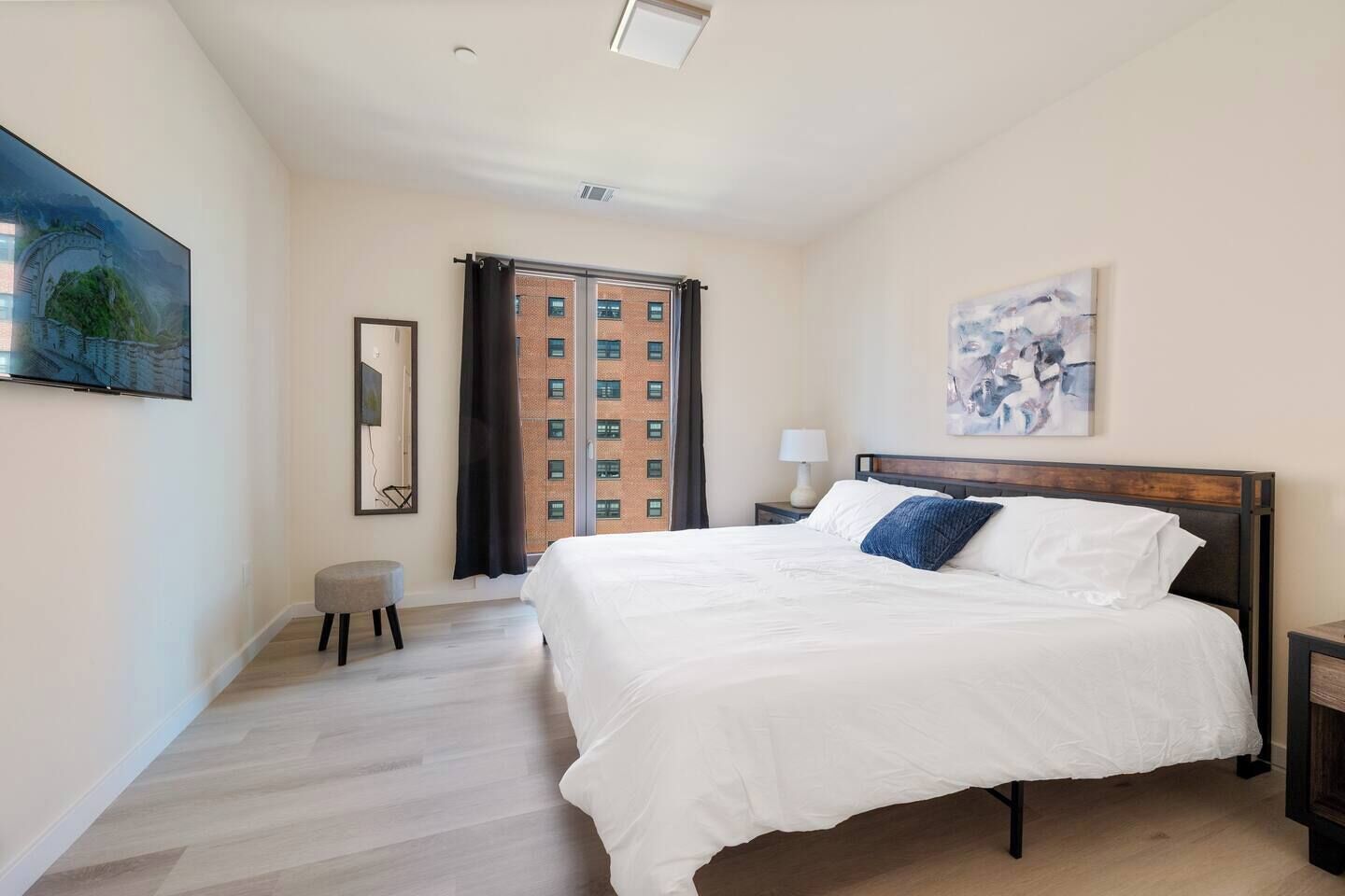 Een slaapkamer met een groot bed, een nachtkastje, een stoel, een raam met uitzicht op gebouwen, en kunstwerken aan de muren.