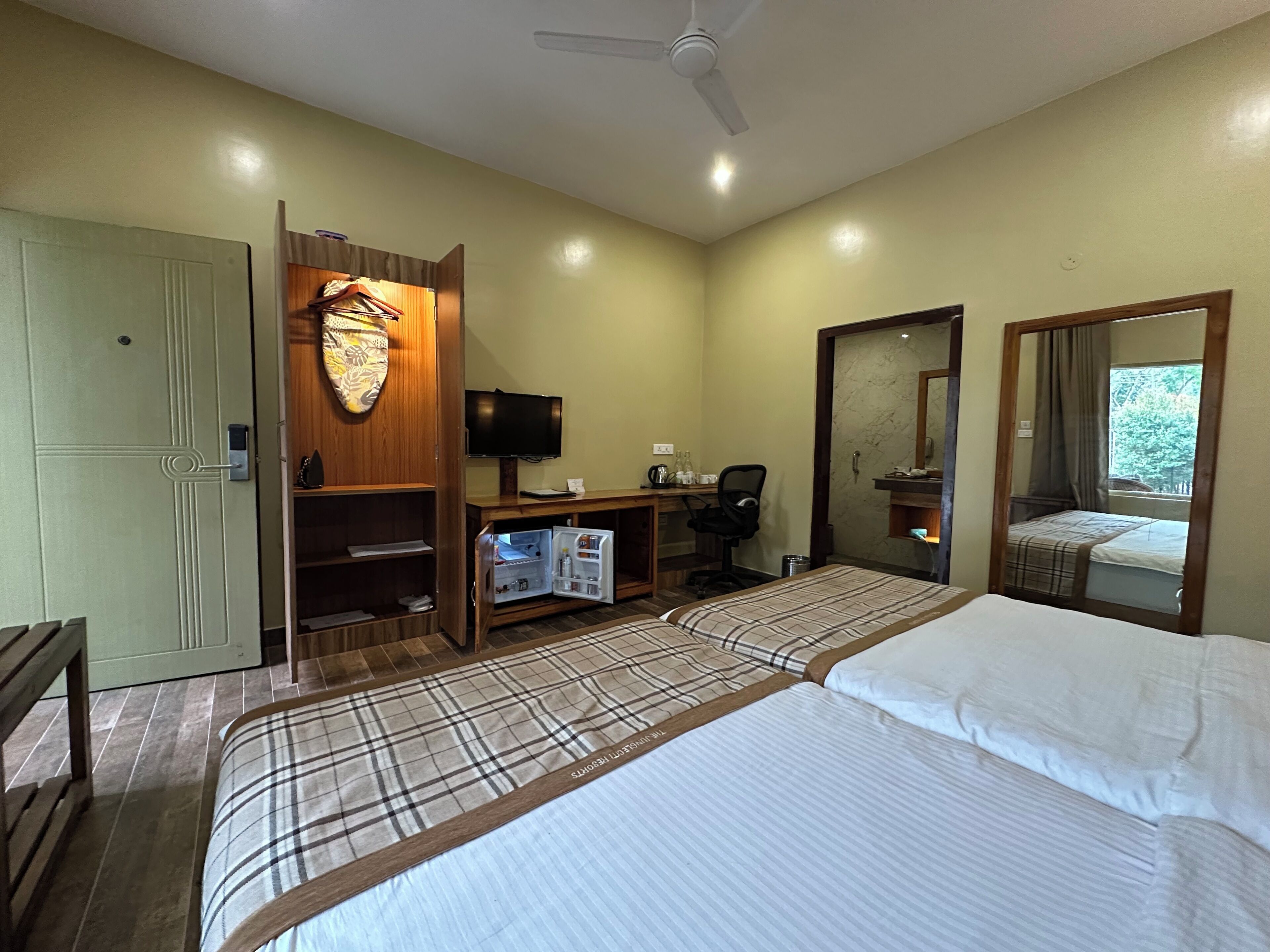 Quarto de hotel com cama, escrivaninha com computador, armário, espelho e ventoinha no teto.