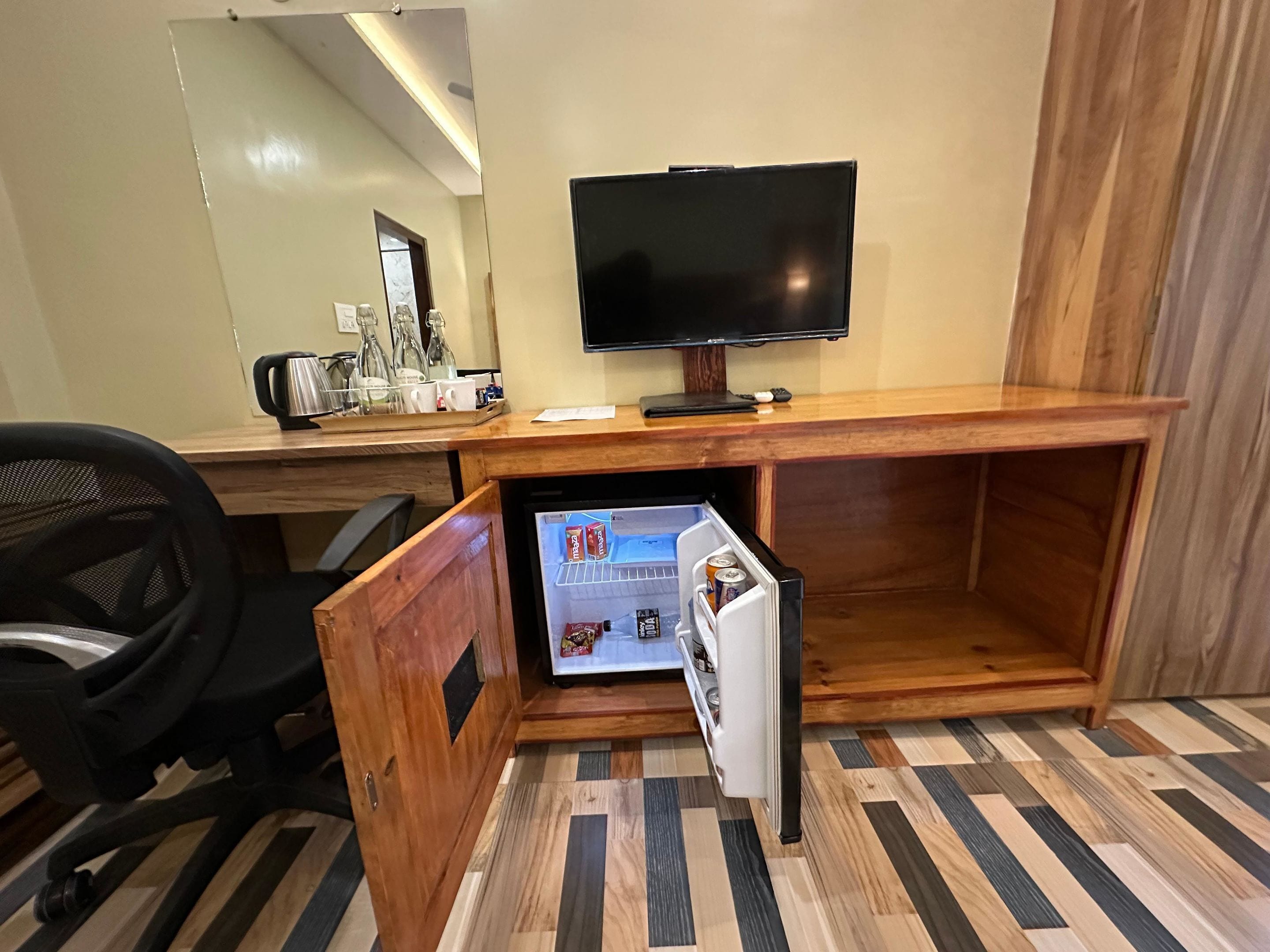 A compact hotel room with a wooden desk, a television, a mini-fridge, and a chair.