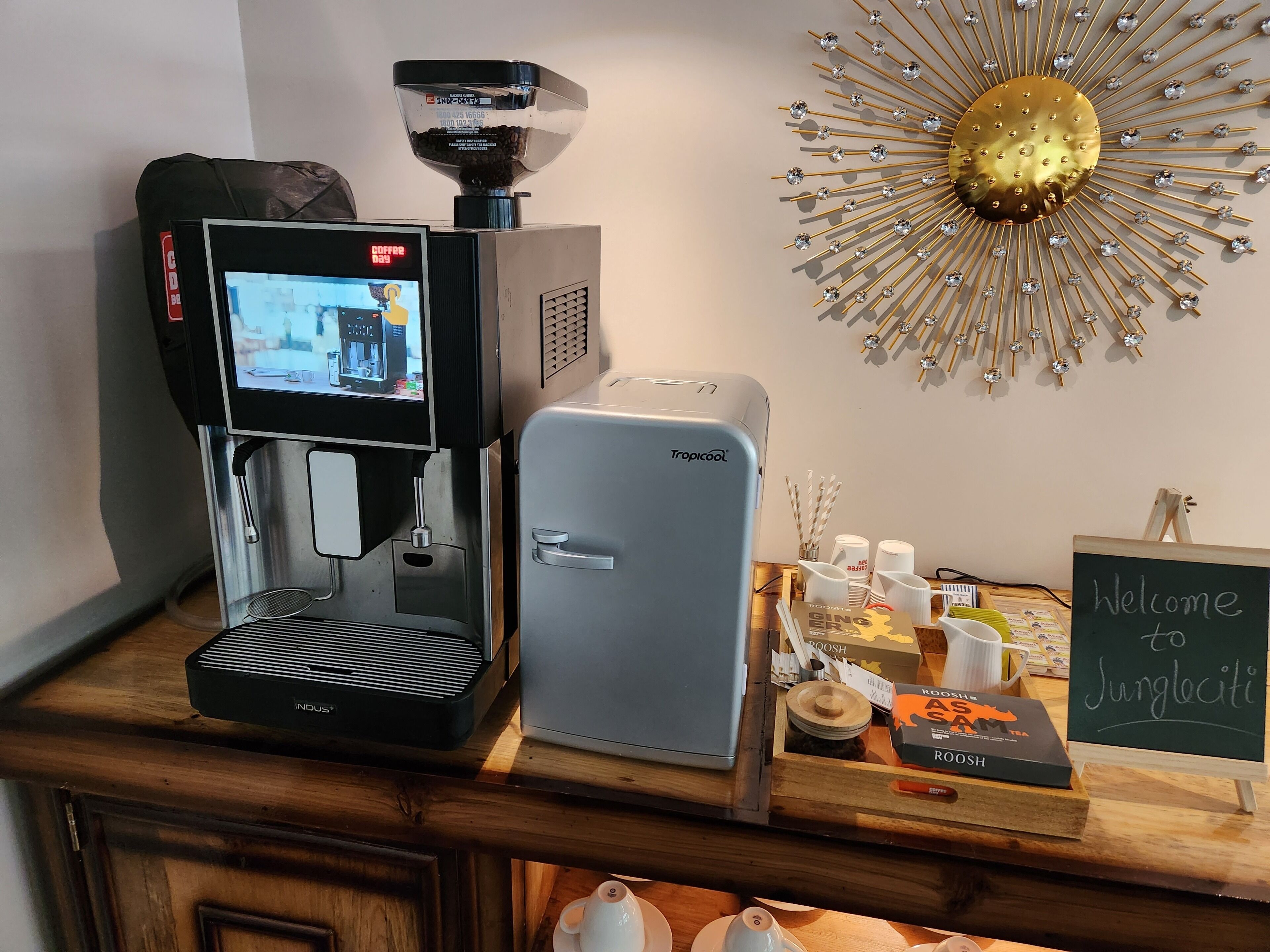 Una cafetera y un refrigerador pequeño sobre una encimera de madera, con un molinillo de café, tazas y un cartel de bienvenida.