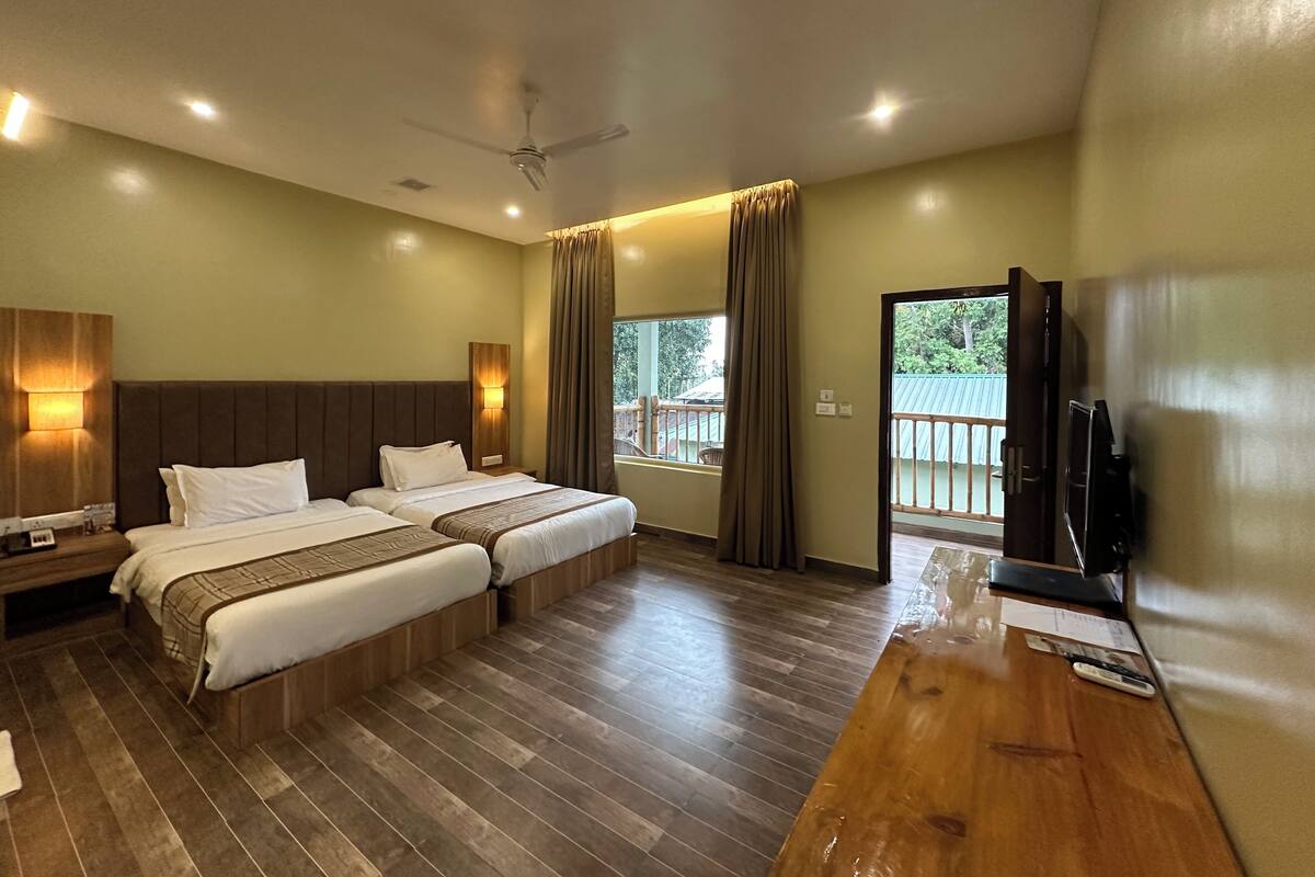 A hotel room with two beds, a wooden desk, a television, and a balcony with a view of greenery.