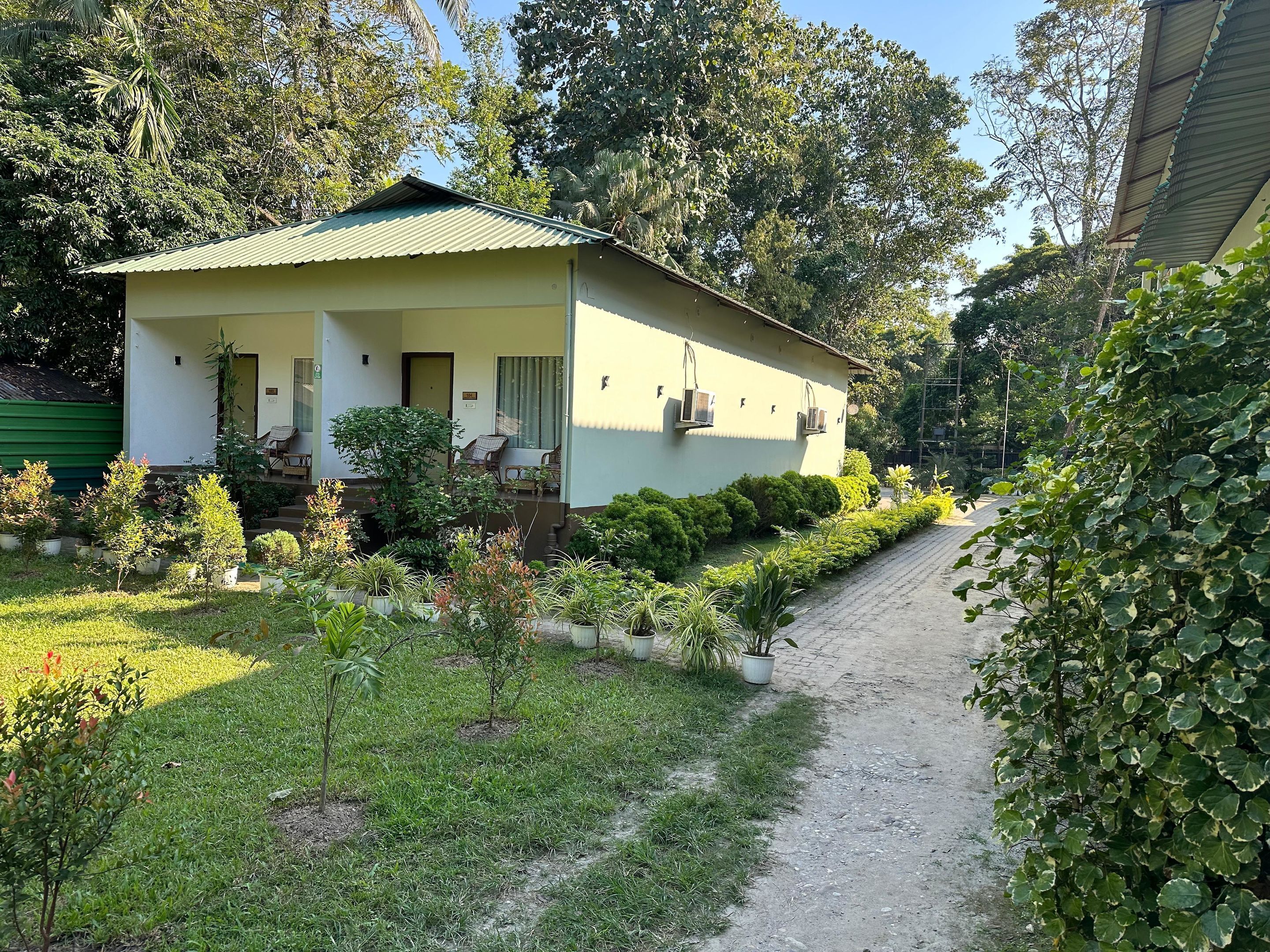 Una casa a un piano con tetto verde, un portico con due sedie e un piccolo giardino con diverse piante e un vialetto lastricato.