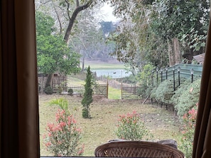 A view from a window overlooking a garden with a wooden fence, a small tree, and a body of water in the distance. - Jungleciti House Kaziranga National Park (Kaliabar)