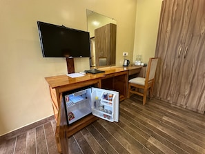 A compact hotel room with a wooden desk, a television, a mini-fridge, a chair, and a wardrobe. - Jungleciti House Kaziranga National Park (Kaliabar)