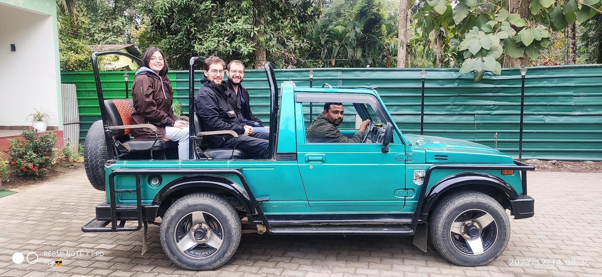Ein türkisfarbenes Pickup-Fahrzeug mit vier Personen, geparkt auf einer befestigten Fläche, im Hintergrund ein grüner Zaun und Vegetation.