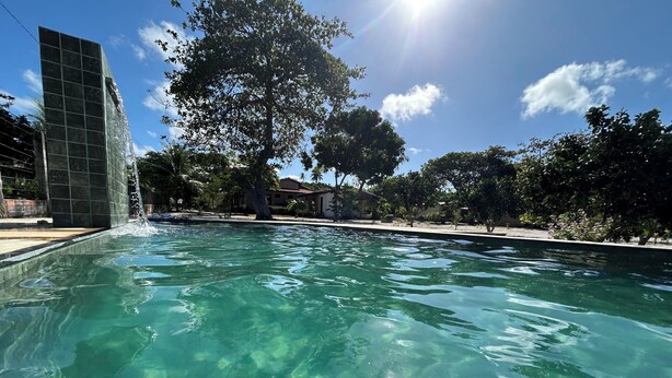 Piscina cristalina con caída de agua, rodeada de árboles y un edificio.