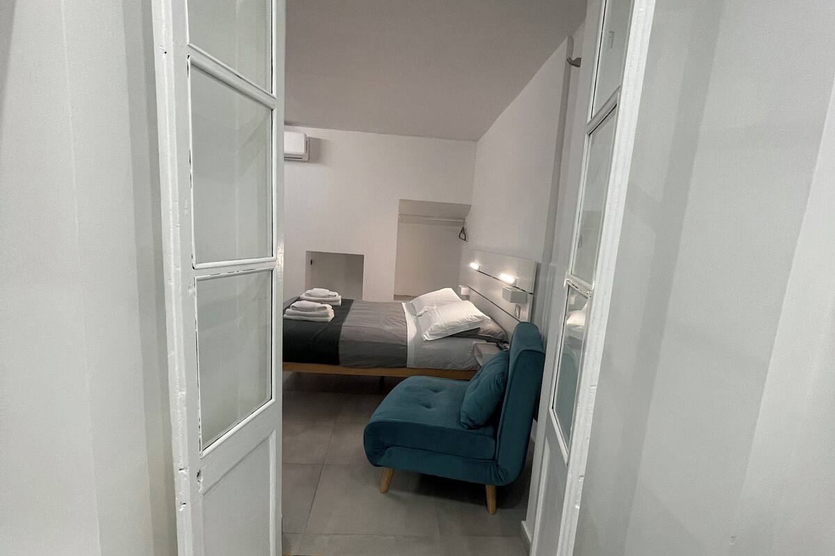 A narrow hallway leading to a bedroom with a bed, a chair, and a bedside table.