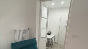 A small, neatly arranged room with a teal-colored bed, a white radiator, a small table with a kettle, and a door leading to another room. - Porta Nuova Suite 57  (Turin)