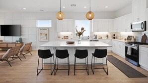 A modern kitchen with white cabinets, a central island with bar stools, a flat-screen TV, and pendant lighting. - Living the DREAM!  Golf and Sunset VIEWS! Brand NEW Home!  Gallery, Indian Palms (Indio)