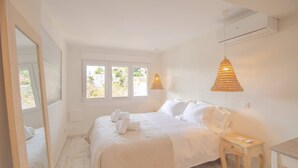 A bedroom with a large bed, two bedside tables, a mirror, and a window with a view of greenery.