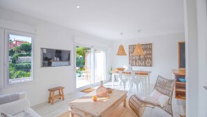 A bright living room with a sofa, coffee table, and wicker chair. There is a dining area with a table and chairs, and a bookshelf with patterned decor.