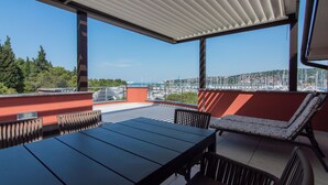 Un espace extérieur aménagé, abrité et doté de tables et de chaises, offrant une vue sur une marina avec des bateaux et sur le paysage urbain au loin.