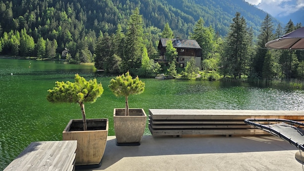 A lakeside cabin with a wooden deck, surrounded by dense forest and mountains.