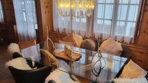 A cozy dining area with a glass table surrounded by black chairs with white fluffy cushions. A chandelier hangs above, and the room has wooden walls and large windows with sheer curtains.