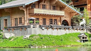 A wooden chalet with a balcony, situated by a lake with a forested backdrop.