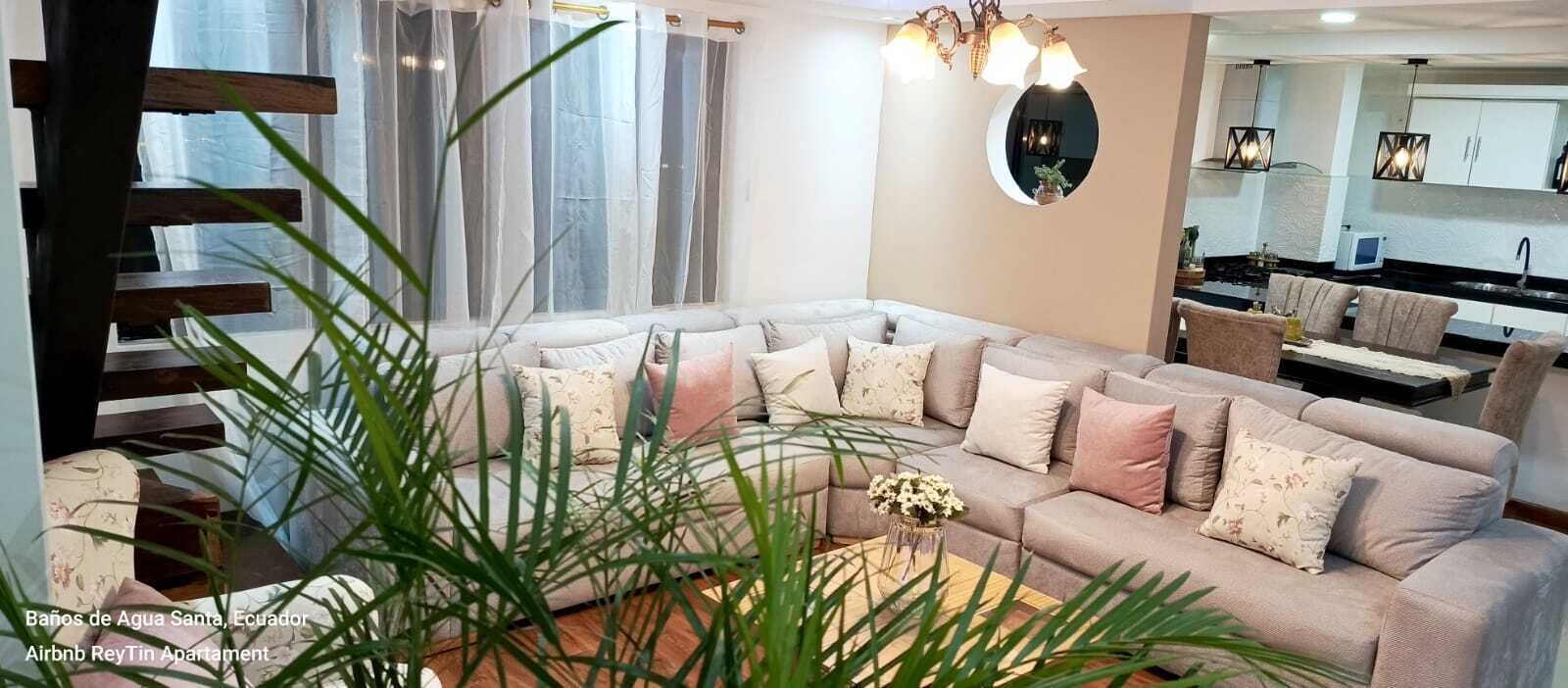 A modern living room with a large L-shaped sofa, a coffee table, and a dining area with chairs. There is a staircase with wooden steps and a plant in the foreground.
