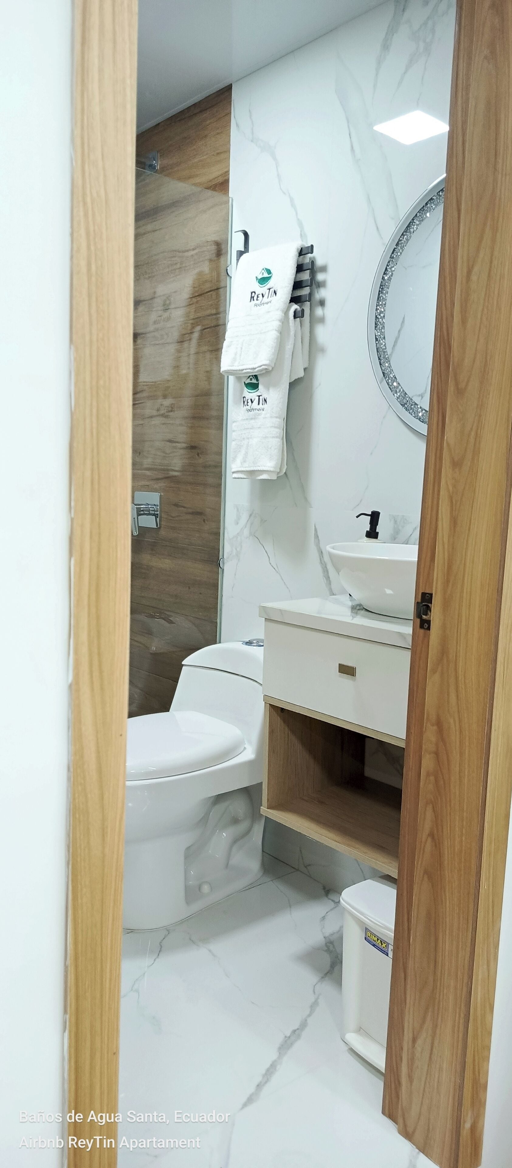 Un baño con inodoro, lavabo y espejo. Toallas colgadas en un perchero. El suelo tiene un diseño de mármol.