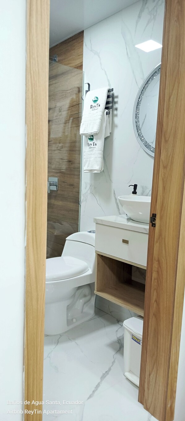 A bathroom with a toilet, sink, and mirror. There are towels hanging on a rack. The floor has a marble pattern.