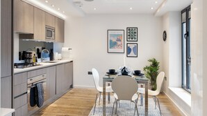 A modern kitchen with wooden cabinets, a microwave, and a dining area with a glass table and white chairs. There are framed pictures on the wall and a potted plant.