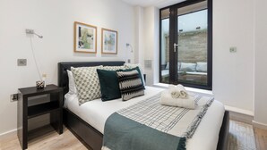 A modern bedroom with a large bed, dark headboard, and a view of a garden through a sliding glass door.